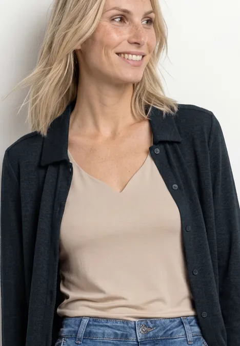 Cheveux blonds aux épaules, top beige à encolure en V et gilet noir sur fond clair.