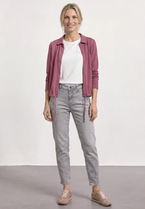 Personne en t-shirt blanc, chemise rose à carreaux, jean gris et baskets roses, posant devant un mur blanc.