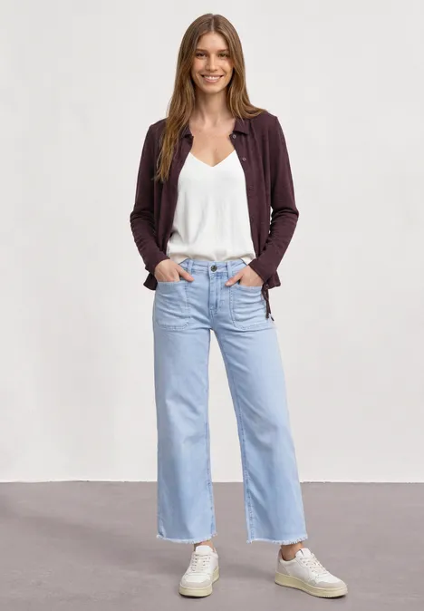 Une femme posant devant un fond neutre porte un débardeur blanc, un jean large bleu clair, un gilet et des baskets.