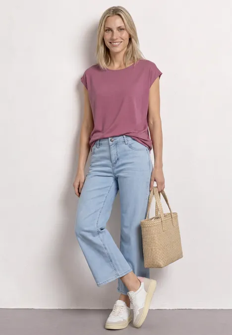 Dropped Shoulder Shirt mit U-Boot-Ausschnitt dark clouded blush Frau in lila Oberteil, hellblauen Jeans, weißen Sneakers und Basttasche.