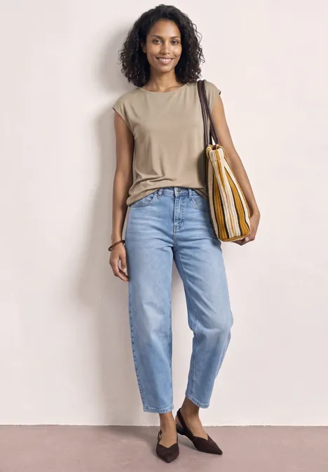Frau steht vor neutralem Hintergrund, trägt beige Bluse, weite blaue Jeans, spitze flache Schuhe und gestreifte Tasche.