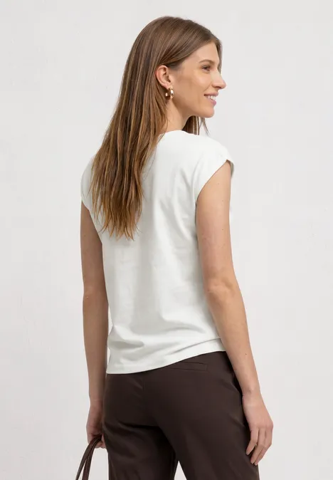 Eine Frau steht seitlich zur Kamera, trägt weißes T-Shirt, braune Hose und hält eine Tasche.