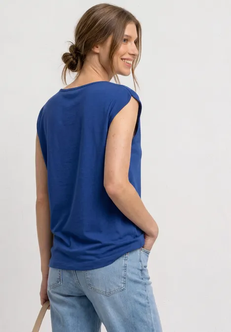 Frau von hinten in blauem ärmellosem Oberteil und heller Jeans, Hand in Tasche, Haare locker zusammengebunden.