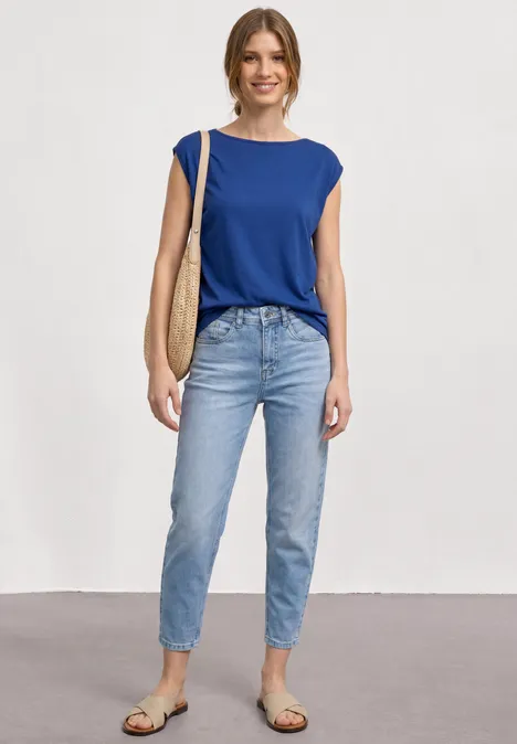 Frau in blauem ärmellosem Oberteil, hellblauen Jeans, Sandalen und Strohtasche vor neutralem Hintergrund.