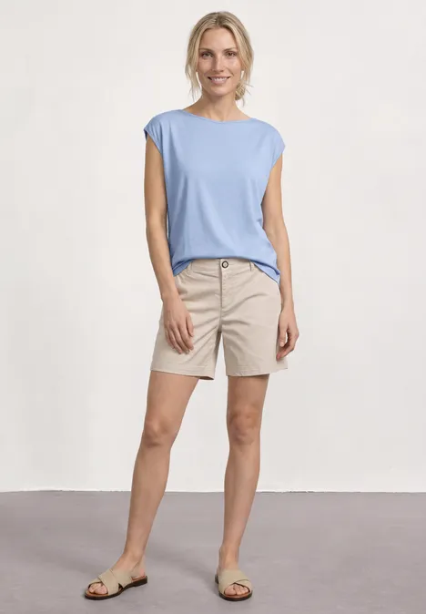 Basic Shirt mit U-Boot-Ausschnitt cashmere blue Frau in hellblauem Top und beigen Shorts, steht vor neutralem Hintergrund.