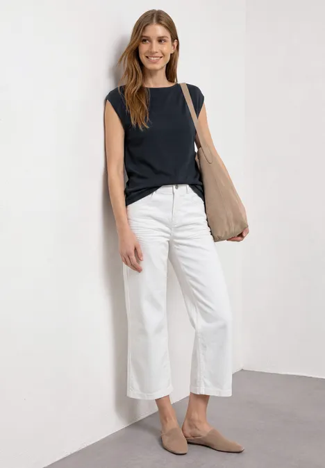 Basic Shirt mit U-Boot-Ausschnitt shadowed navy Frau lehnt an weißer Wand in schwarzem Top, weißen Culottes, beigen Slippern und beiger Tasche.