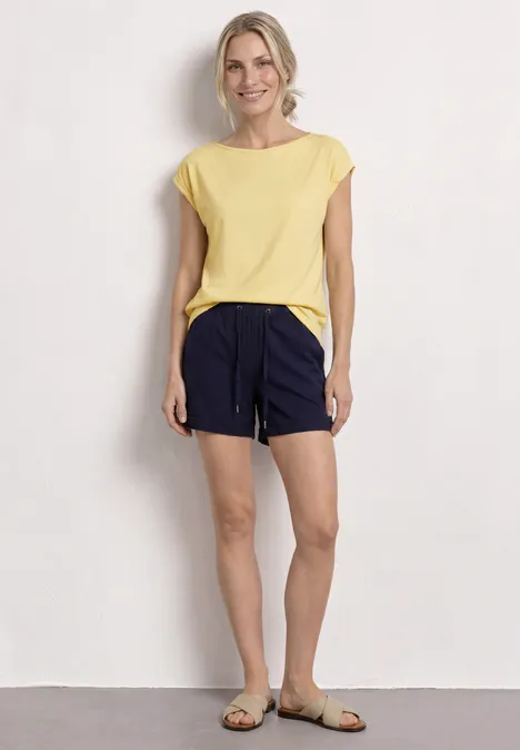 Basic Shirt mit U-Boot-Ausschnitt soft yellow Eine Frau trägt ein hellgelbes T-Shirt, dunkelblaue Shorts mit Kordelzug und sandfarbene Sandalen vor weißer Wand.