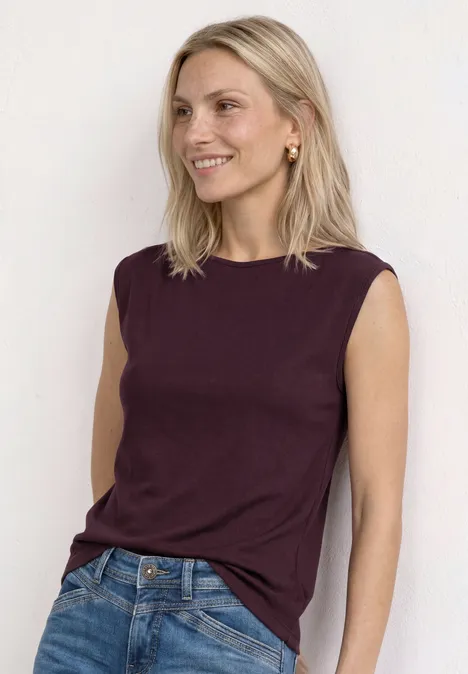 Basic Shirt mit U-Boot-Ausschnitt rustic rouge Frau trägt ärmelloses bordeauxfarbenes Top und blaue Jeans, lehnt an weißer Wand.