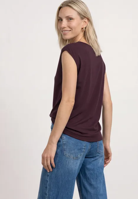 Basic Shirt mit U-Boot-Ausschnitt rustic rouge Eine Person mit blondem Haar trägt ein ärmelloses dunkelviolettes Top und blaue Jeans und blickt nach hinten.