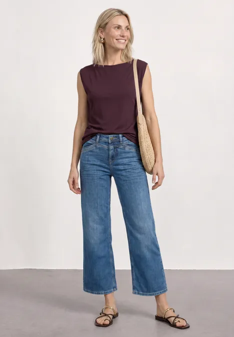 Basic Shirt mit U-Boot-Ausschnitt rustic rouge Frau vor neutralem Hintergrund in lila Top, weitem Jeans, flachen schwarzen Sandalen und Korbtasche.