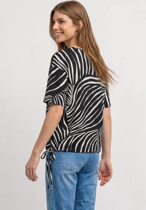 Vrouw draagt zwart shirt met wit zebraprint, halflange mouwen en zijstrik, gecombineerd met blauwe jeans.