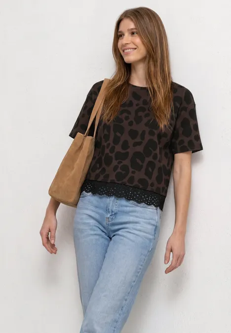 Vrouw voor witte achtergrond met een zwart shirt met print, lichte jeans en een bruine tas.