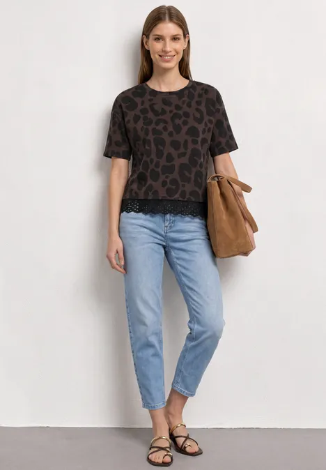 Vrouw in luipaardprint t-shirt, lichtblauwe spijkerbroek, zwarte sandalen en bruine tas voor neutrale muur.