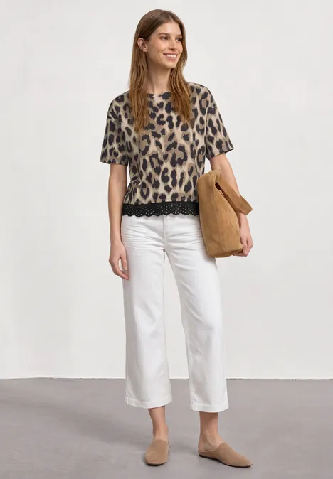 Vrouw met luipaardprintshirt, witte culotte, beige slippers en bijpassende tas tegen neutrale achtergrond.