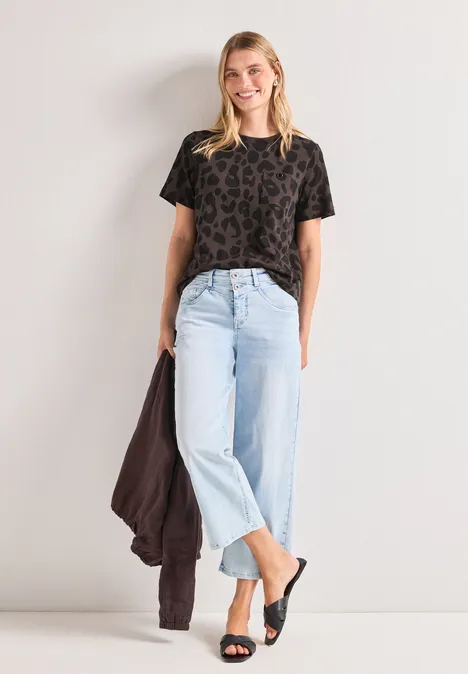 Frau in gemustertem Shirt, hellen Jeans und schwarzen Sandalen lehnt entspannt an der Wand, Jacke in der Hand.