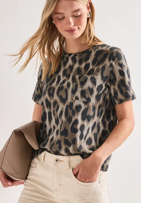 Frau in legerem Leoparden-Print-Shirt hält eine beige Clutch, lässig in Hose gesteckt.