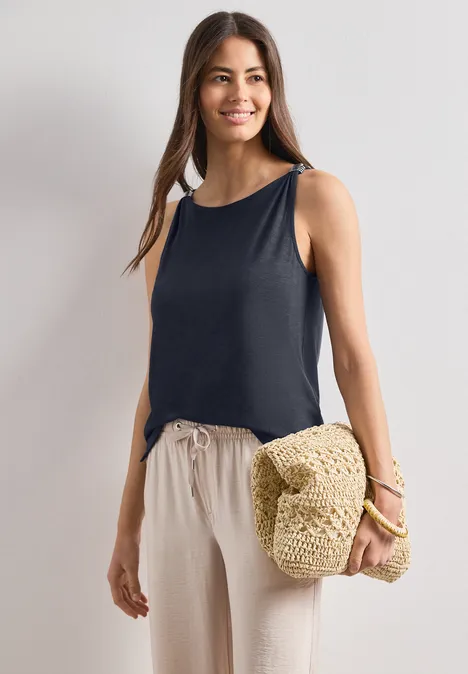 Eine Frau trägt ein ärmelloses schwarzes Top, bequeme helle Hosen und hält eine Strohtasche.
