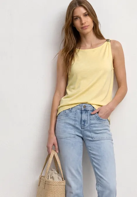 Frau vor neutralem Hintergrund trägt gelbes Top, blaue Jeans und hält Tasche.