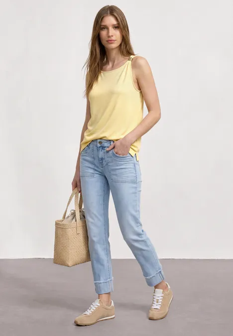 Frau trägt gelbes Top, blaue Jeans, beige Sneaker und Strohtasche vor neutralem Hintergrund.
