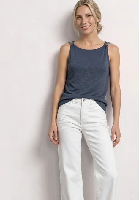Top mit U-Boot-Ausschnitt und Schulterdetail ashen blue Frau in blauem ärmellosen Top und weißen Jeans steht vor neutralem Hintergrund.