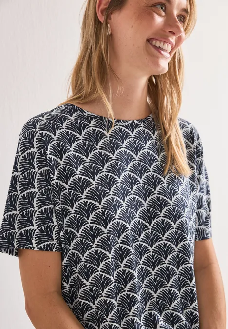 Femme aux cheveux blonds porte un haut manches courtes noir orné d’un motif blanc en forme de feuilles.