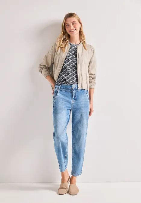 Femme en veste beige, T-shirt à motif, jean bleu clair et mocassins beiges devant un fond neutre.