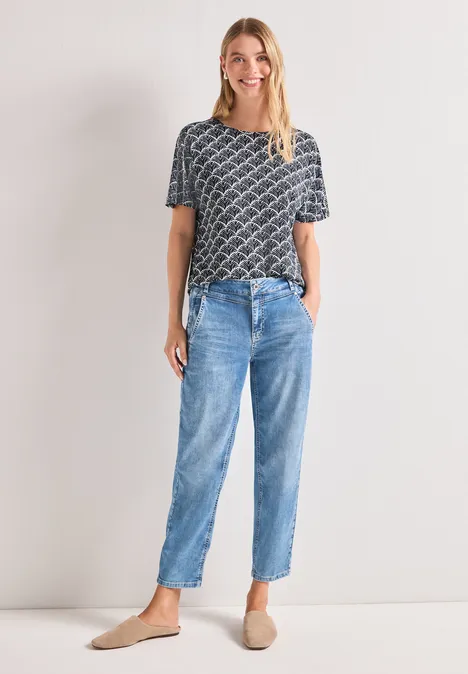 Femme portant un t-shirt gris à motif, des jeans bleus et des mules beiges sur fond clair.