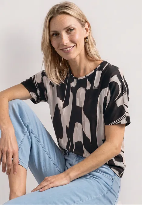 Vrouw zit met gebogen been, draagt een zwart-wit dessin top en lichtblauwe jeans.