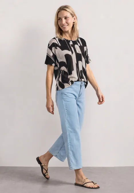 Vrouw in patroonblouse, lichte jeans en zwarte sandalen voor een neutrale achtergrond.