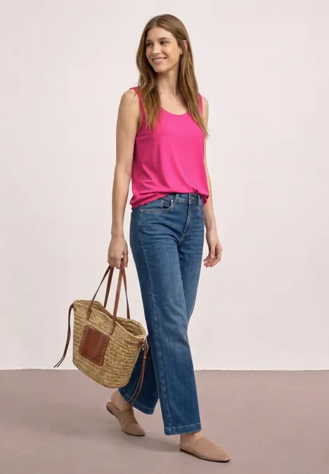 Frau in pinkem Top, blauen Jeans und beigen Schuhen mit Korbtasche vor neutralem Hintergrund.