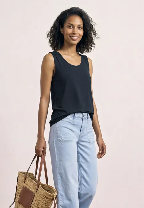 Frau mit schwarzem Top, hellblauen Jeans und geflochtener Tasche vor hellem Hintergrund.