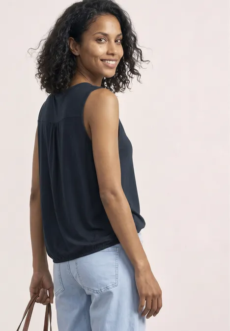 Frau in schwarzem ärmellosen Top und hellblauen Jeans, hält zum seitlichen Blick eine Handtasche.