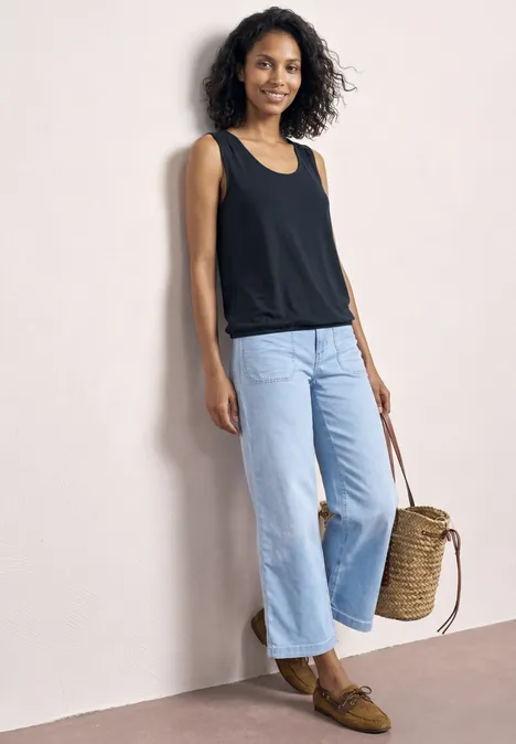 Frau lehnt an heller Wand, trägt schwarzes Top, weite helle Jeans, braune Loafers und hält einen geflochtenen Korb.