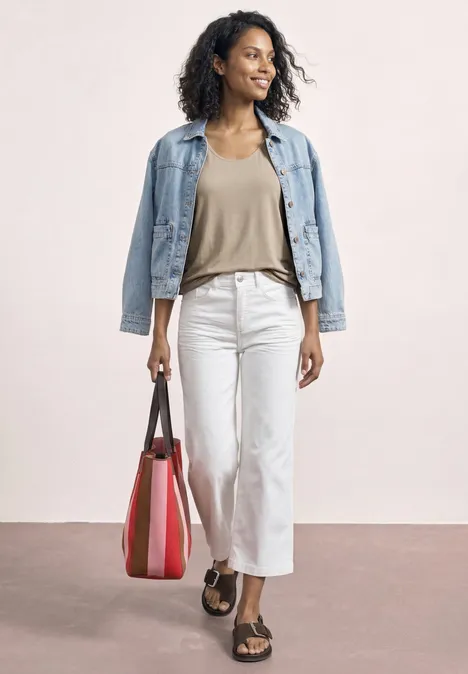 Top aus Jersey mit Elastiksaum sepia sand Frau mit Jeansjacke, beiger Bluse, weißen Hosen, Sandalen und roter Tasche vor neutralem Hintergrund.