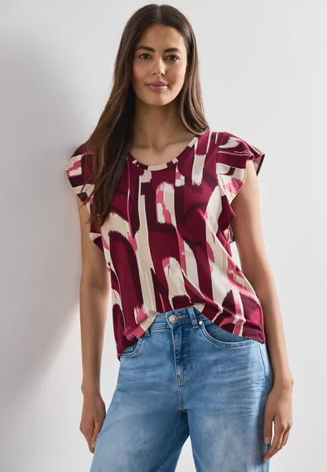 Vrouw draagt een bordeaux-rode top met witte print en blauwe jeans.