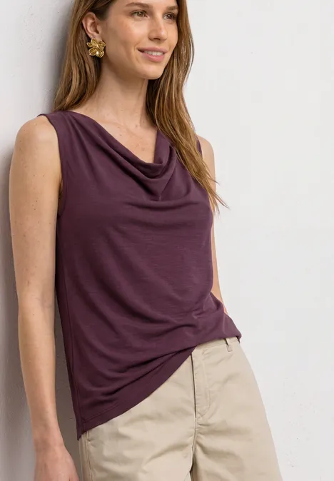 Vrouw met paarse mouwloze draperingstop en lichte broek voor een neutrale achtergrond.