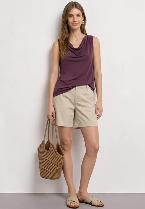 Vrouw draagt mouwloze paarse top, beige short, platte sandalen en gevlochten strozak.