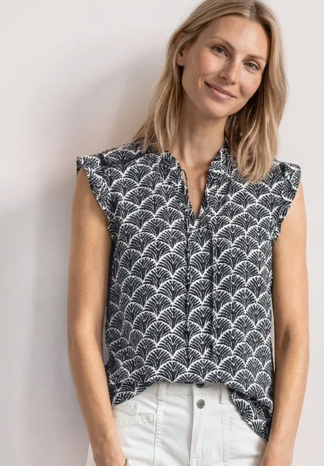 Dame trägt ärmellose Bluse mit geometrischem Muster, vorn leicht in die Hose gesteckt.
