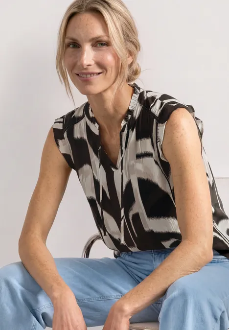 Eine sitzende Person mit schwarz-weiß gemusterter, ärmelloser Bluse und hellblauen Jeans.