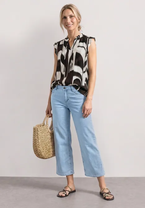 Frau in ärmelloser schwarz-weißem Print-Bluse, hellen, gekürzten Jeans, schwarzen Sandalen und Korbtasche.