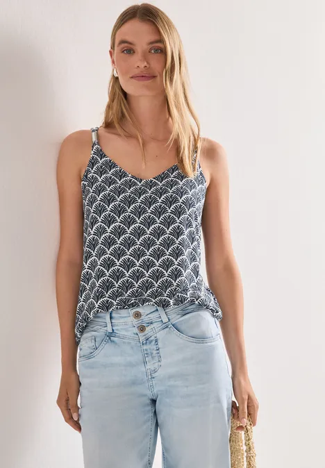 Frau trägt gemustertes Top, helle Jeans und hält eine Strohtasche vor weißem Hintergrund.