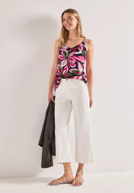 Femme en top sans manches à imprimé coloré, pantalon blanc large, sandales fines blanches, tenant une veste noire.