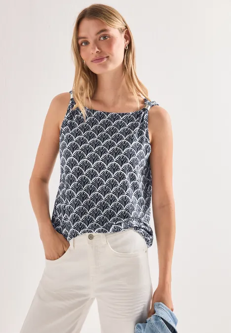 Femme portant un top bleu à motifs et un jean blanc devant un fond clair.