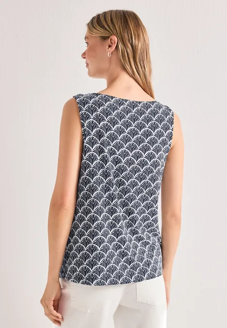 Femme de dos portant un top sans manches à motifs éventails bleu-blanc et un pantalon blanc sur fond neutre.