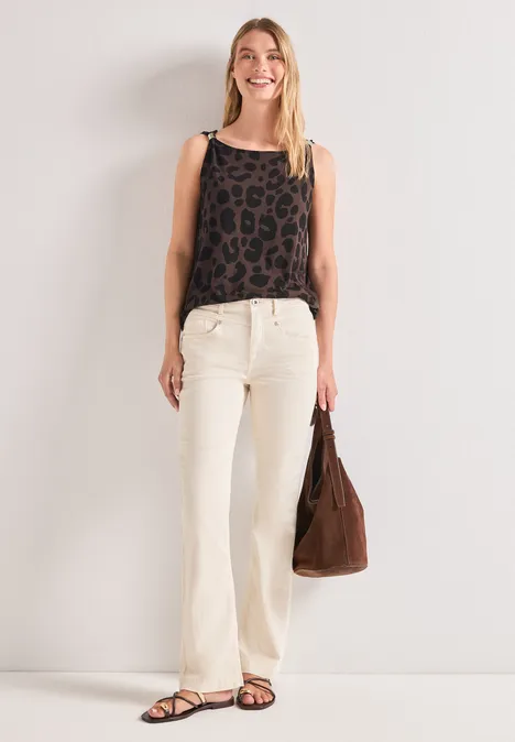Frau in weißen Schlagjeans und Leoparden-Top, hält eine braune Tasche vor weißer Wand.