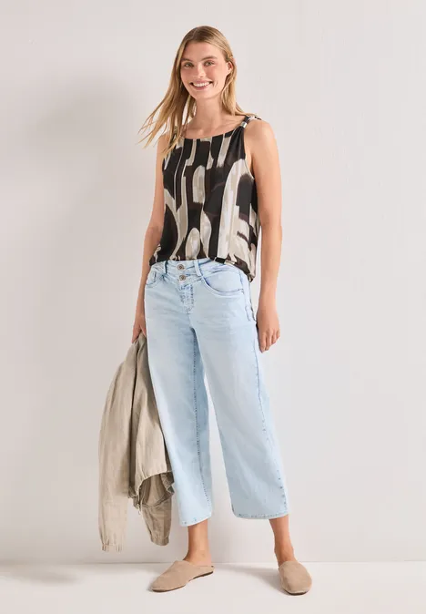 Frau in gemustertem Top, weiten hellblauen Jeans und flachen Schuhen hält eine Jacke in der Hand.