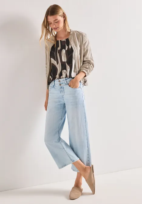 Frau in beiger Jacke, gemustertem Top, weiten hellblauen Jeans und beigen Slippern vor hellem Hintergrund.