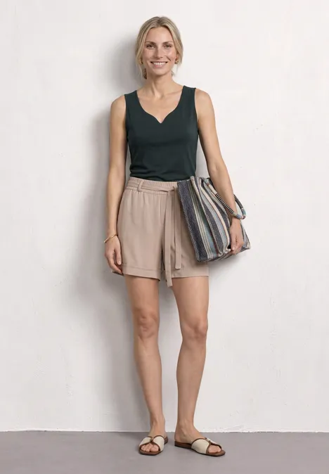 Eine schlanke Frau in dunkelgrünem Top, beigen Shorts und Sandalen hält eine gestreifte Tasche.