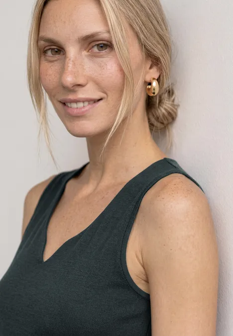 Eine blonde Frau mit Dutt, goldenen Creolen und ärmellosem Oberteil lächelt in die Kamera.