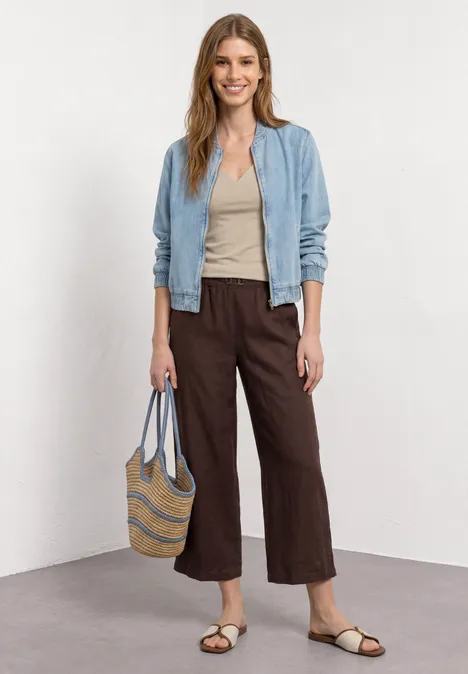 Frau in blauer Jacke, beigem Top, brauner Culotte, Sandalen und Korbtasche vor hellem Hintergrund.